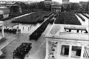 Schwarzweißfoto eines großen Platzes, der von antikisierenden Gebäuden gerahmt wird. Auf dem Platz stehen präzise geordnet große Gruppen von Menschen