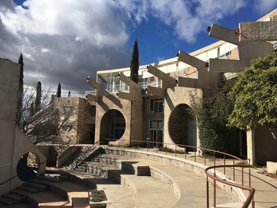 Ansicht eines Häuserrondells in Arcosanti. Ein Stufenrondell im Stil eines Amphiteaters, das von Gebäuden gerahmt wird. Die Fenster der Gebäude sind in vorgezogenen Runden Wandaussschnitten. Zwischen dem Erdgeschoß und dem Obergeschoß gliedern weit hervorgezogene Trägerstrukturen aus Beton in V-Form die Fassade.