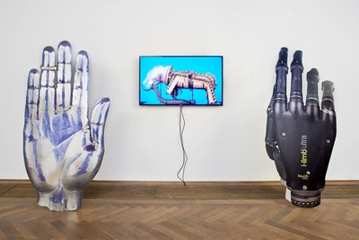 Vor einer weißen Wand stehen zwei große mechanische Hände. Zwischen ihnen hängt ein Monitor.