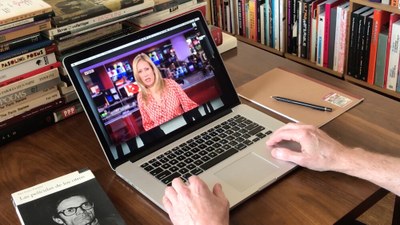 Eingeschalteter Laptop auf Schreibtisch. Zwei Hände bedienen hin. Im Hintergund befinden sich viele Bücher.