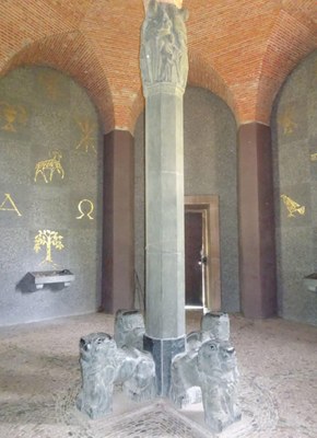 Foto. Kircheninnenraum mit mittiger Säule und goldenen Symbolen an den Wänden.