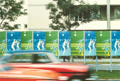 Schnappschuss Straßenszenerie. Rotes Auto vor einer aneinanderreihung von blau-grünen Ankündigungsplakaten für die Olympischen Spiele 1972