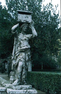 Moosbewachsene Skulptur aus Stein in einem grünen Garten. Sie trägt einen Kasten auf dem Kopf. 