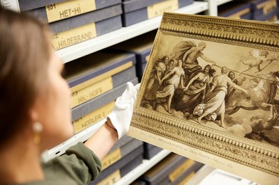 Eine Frau ssteht in einem Archiv und hält die Reproduktion eines prächtigen Deckengemäldes in den Händen.