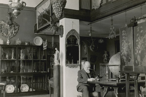 Schwarzweißfoto eines alten Mannes in Anzug in einem großen Innenraum, in dem allerlei Gegenstände herumstehen.