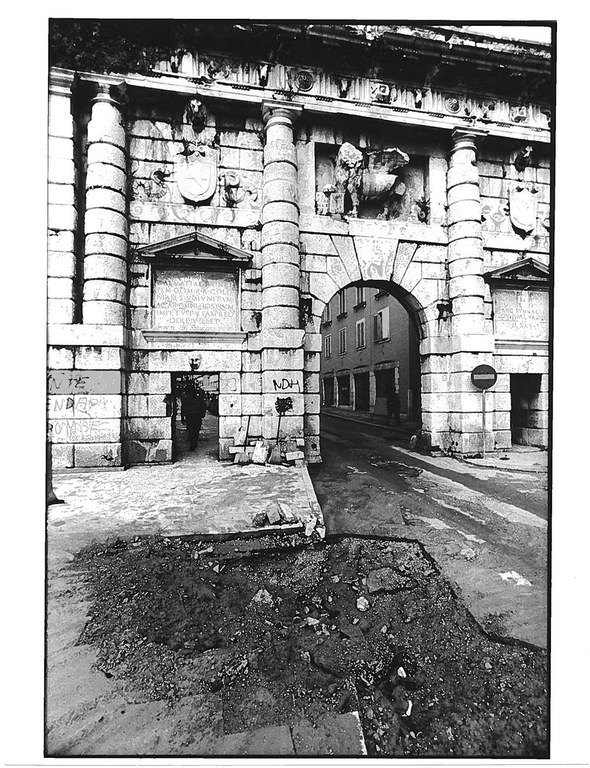 Altes schwarz/weiß Foto. Man sieht einen Toreingang in einer hohen Mauer mit EInschusstellen. DAvor auf der Straße tiefe Löcher