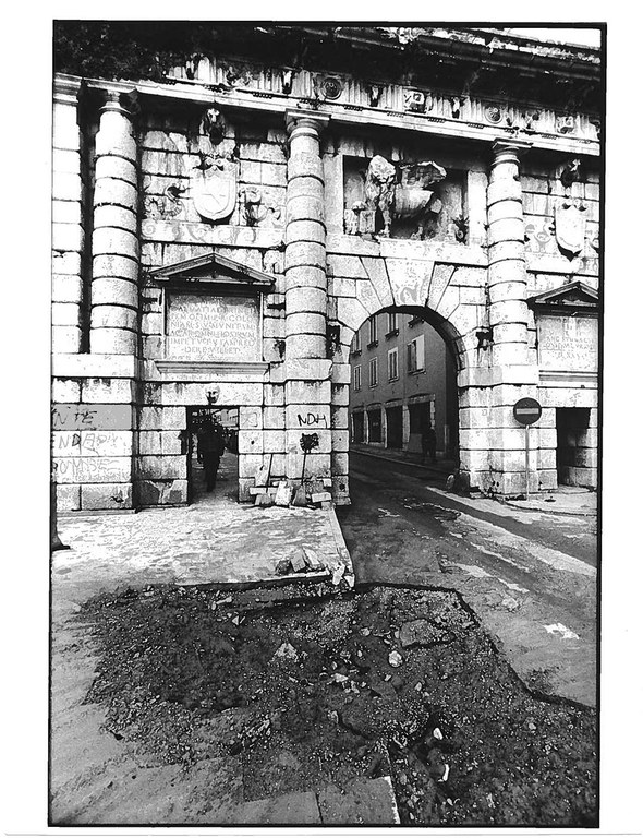 Altes schwarz/weiß Foto. Man sieht einen Toreingang in einer hohen Mauer mit Einschussstellen. Auf der Straße davor sind tiefe Löcher.