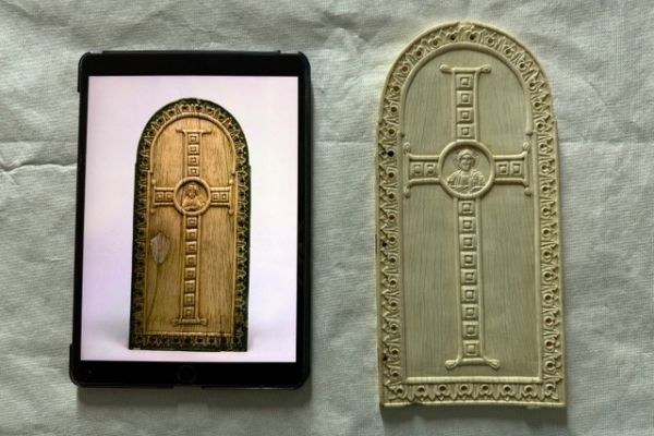 Eine geschnitzte Elfenbeinplatte. Das Motiv ist ein Kreuz in dessen Mitte das Portät eines Mannes liegt. Neben der Platte liegt ein Tablet, auf dem ein Foto einer fast identischen Platte geöffnet ist.