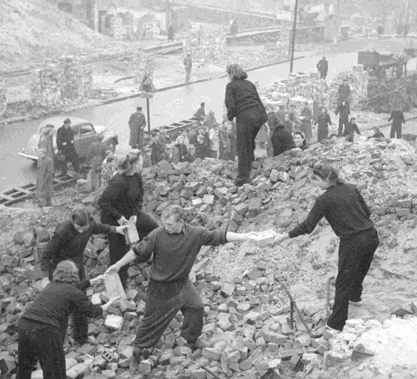 Schwarzweißfoto. Junge Menschen stehen auf dem Bauschutt einer Häuserruine und reichen Backsteine umher.
