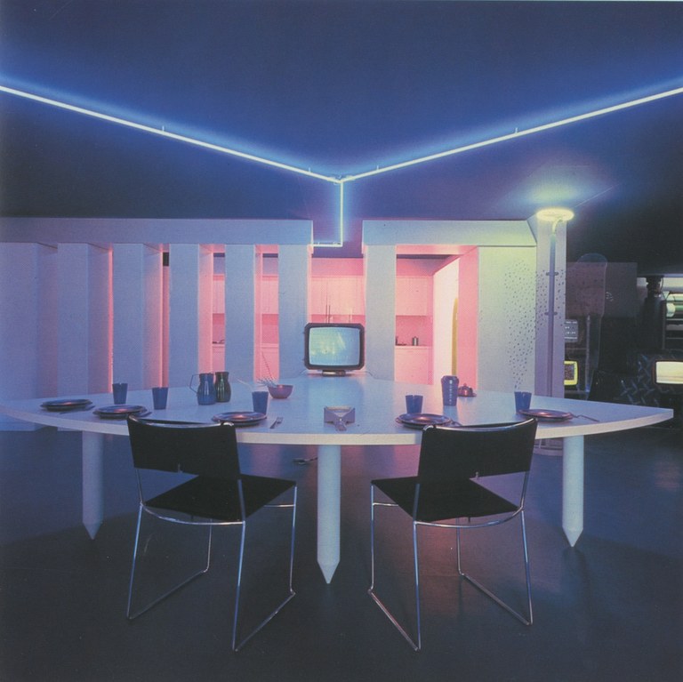 Foto: Futuristisches Interieur. Gedeckter Tisch, ausgerichet auf einen angeschalteten Röhrenfernseher. Beleuchtung durch Neonlicht.