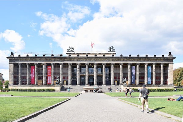 Foto: Flaches Gebäude aus Stein mit vielen Säulen (Frontansicht) vor blauem Himmel. Zu dem Gebäude führt ein breiter Weg mit grünem RAsen links und rechts